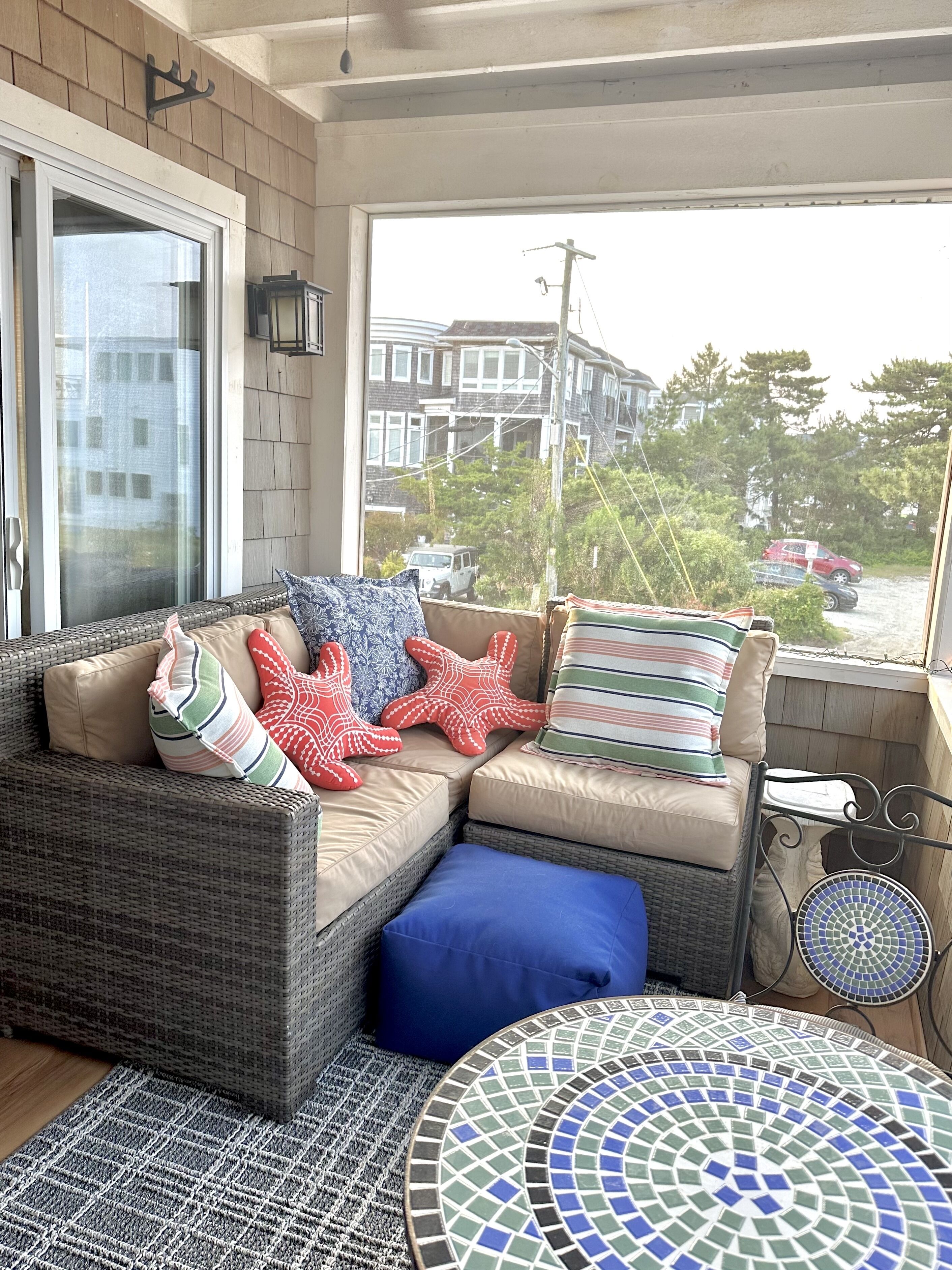 A wicker sofa with cushions, a patterned rug, and a mosaic table in a sunlit room with a view of buildings and trees.