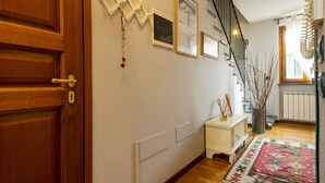 A hallway with a wooden door, a white wall with framed pictures, a small white table with a vase, a staircase, and a patterned rug. - Lovely apartment for 4 guests with A/C, WIFI, TV, terrace and pets allowed (Capannori)