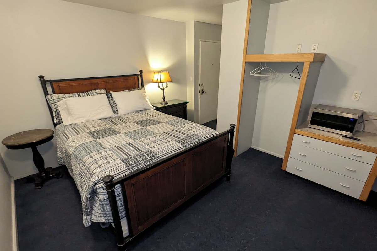 A bedroom with a wooden bed, a bedside table, a lamp, a dresser, and a microwave.