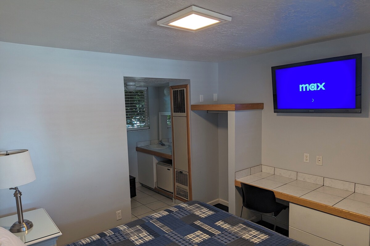 A hotel room with a bed, a desk, a TV, and a bathroom visible through an open door.