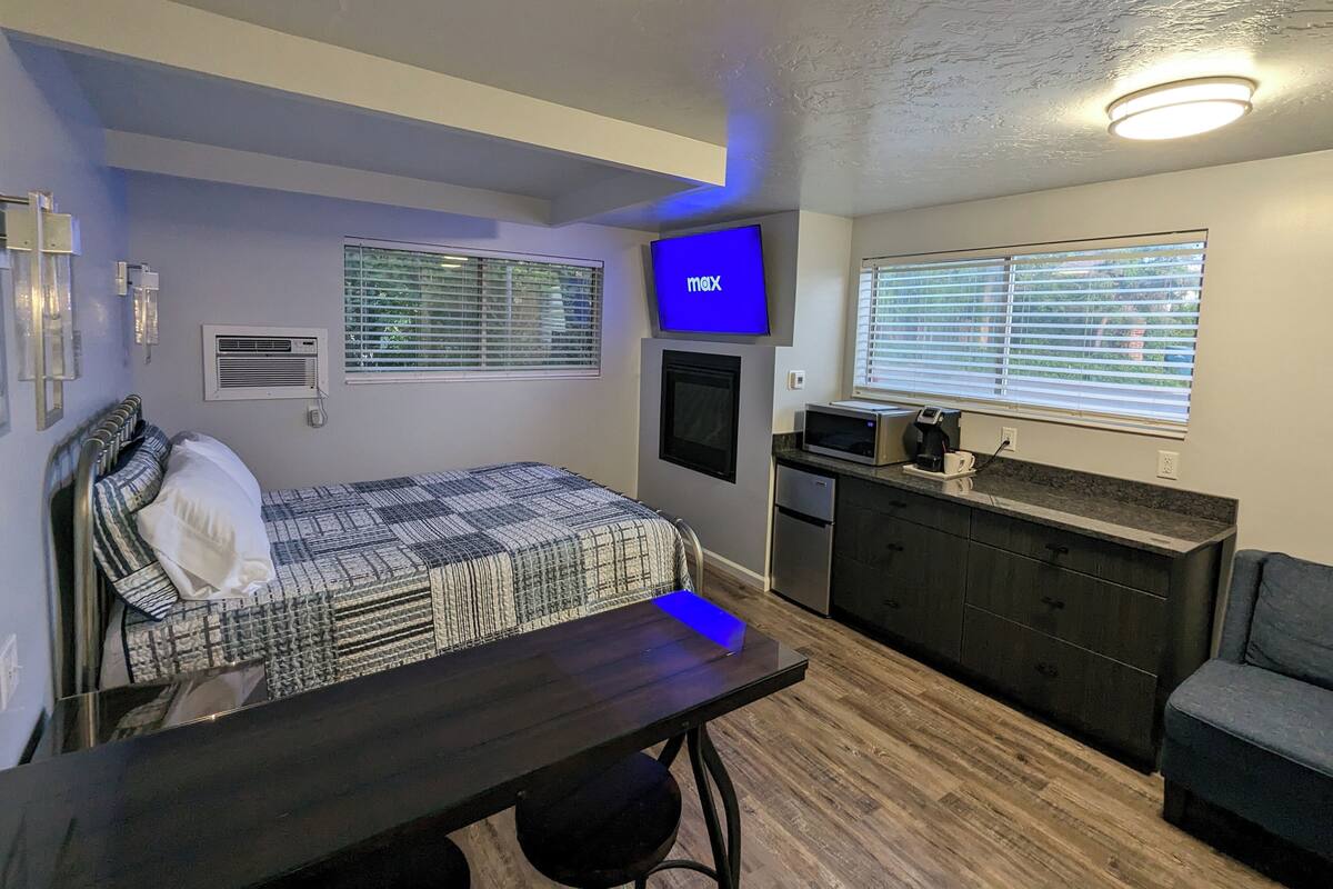 A compact bedroom with a bed, a desk, a TV, a microwave, and a small kitchenette.