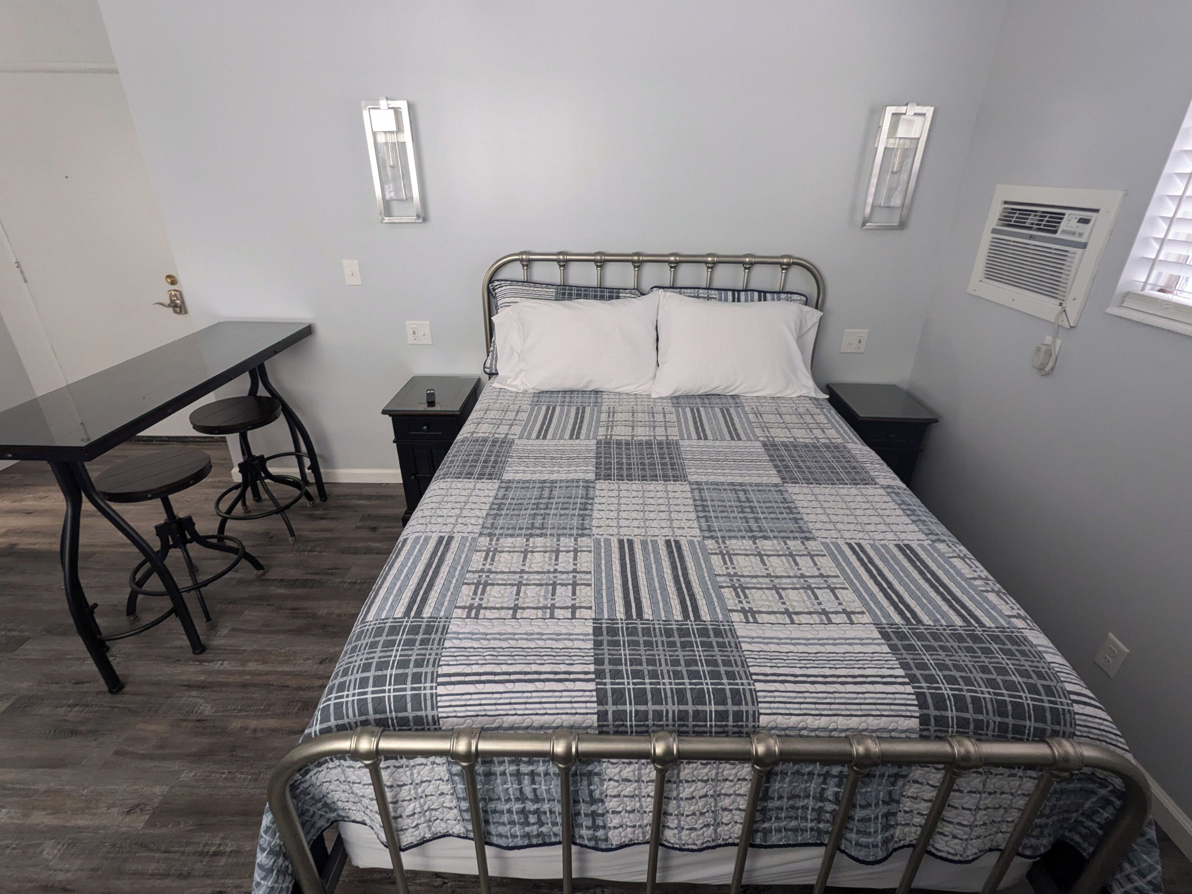 Ein Schlafzimmer mit einem Metallbettgestell, einem Holztisch mit zwei Hocker, einer gemusterten Bettdecke und zwei Nachttischen mit Wandleuchten.