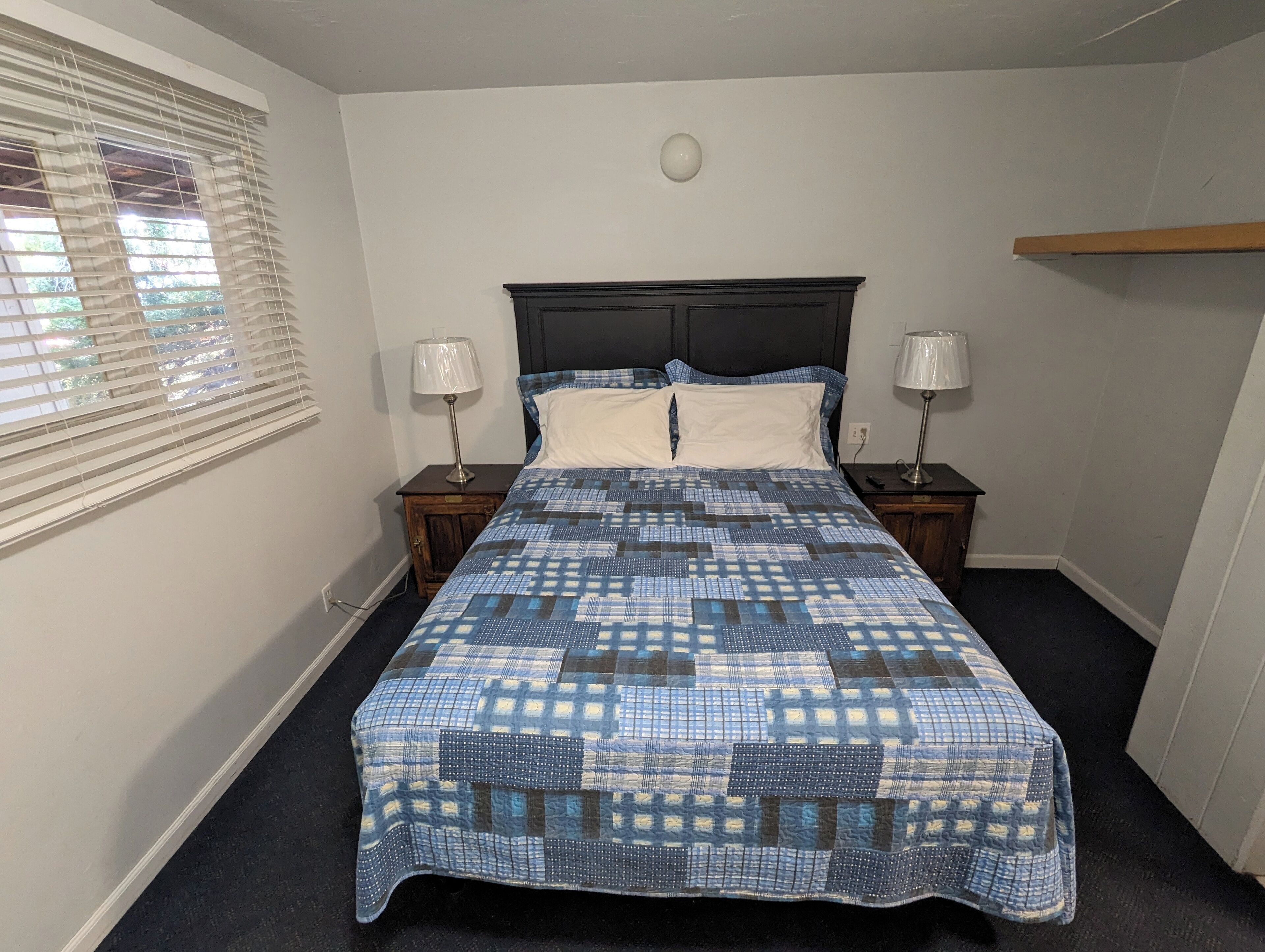 A bedroom with a bed, two bedside tables, a window with blinds, and a wall-mounted light fixture.