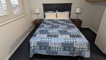 A bedroom with a bed, two bedside tables, a window with blinds, and a wall-mounted light fixture.