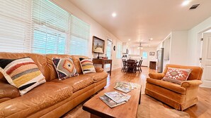 A spacious living room with a brown leather sofa, a wooden coffee table, and a brown armchair. There is a dining area with a wooden table and chairs in the background. - The Whitehouse – Louisiana Country Getaway on 8 Peaceful Acres with Pond Views (Robert)