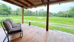 A covered outdoor seating area with a wooden bench and a lantern, overlooking a grassy field and trees. - The Whitehouse – Louisiana Country Getaway on 8 Peaceful Acres with Pond Views (Robert)