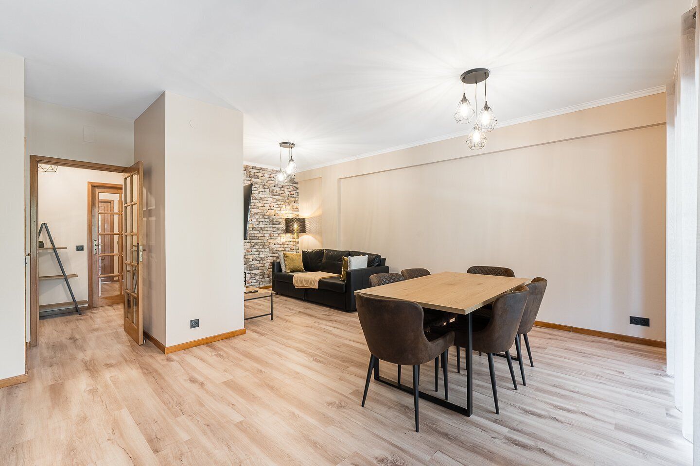 Un salon moderne avec coin repas, doté d’un parquet, d’un mur en briques apparentes et d’un canapé en cuir noir.