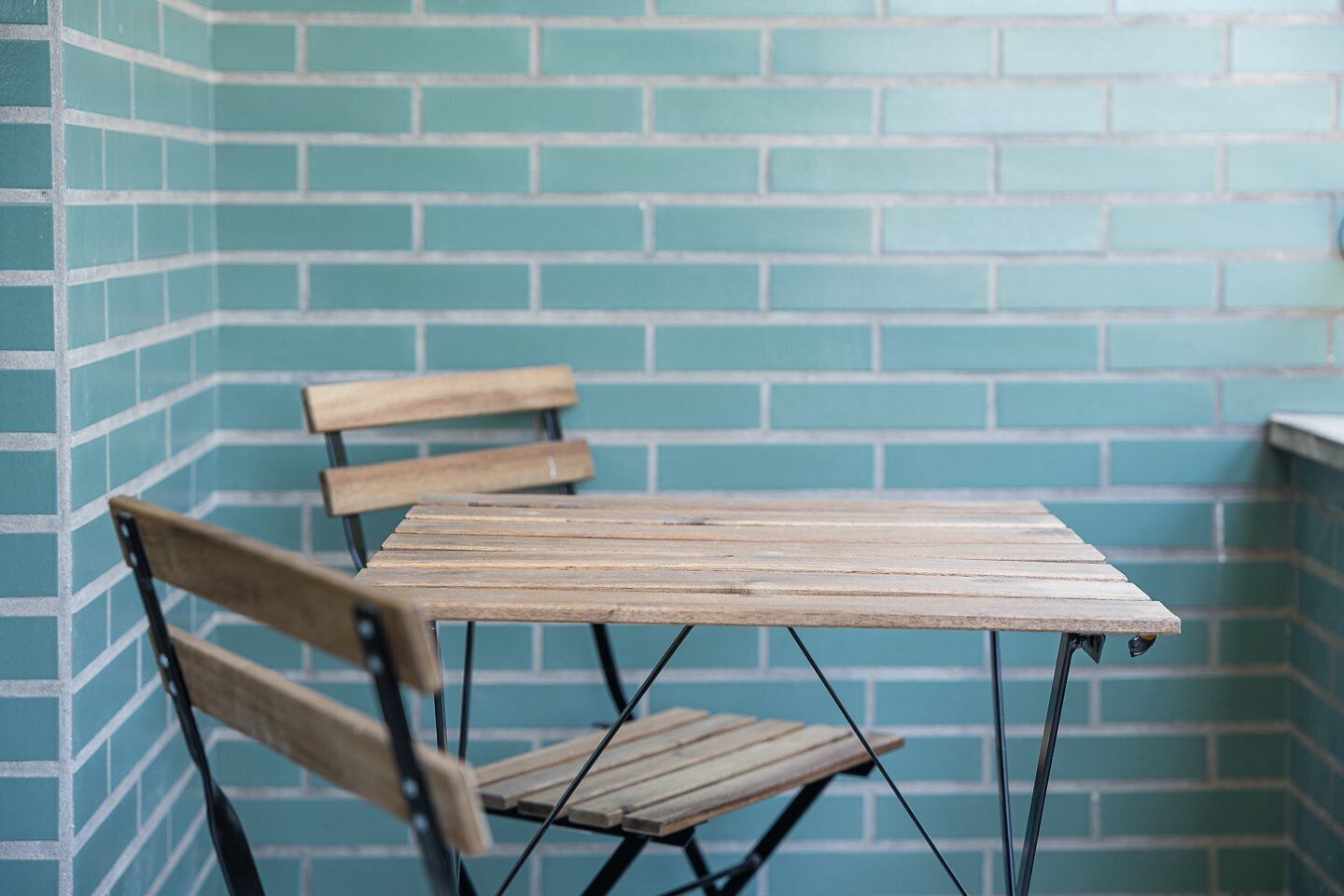 Una pequeña mesa plegable de madera con patas metálicas, acompañada por dos sillas de madera con estructura de metal, frente a una pared de ladrillo color turquesa.
