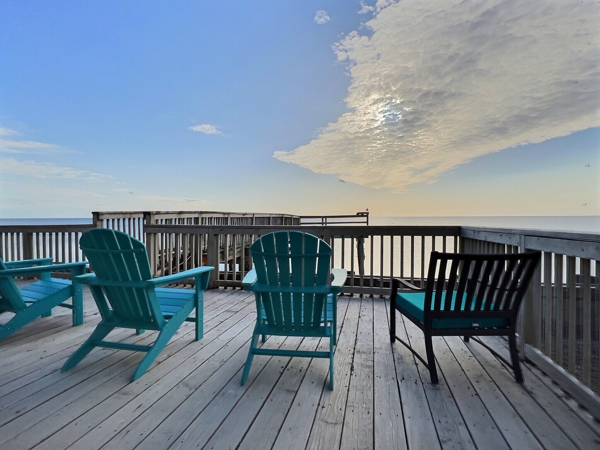 Une terrasse en bois, agrémentée de chaises Adirondack turquoise et d’un banc, qui offre une vue sur une mer calme et une jetée en bois.