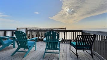 Une terrasse en bois, agrémentée de chaises Adirondack turquoise et d’un banc, qui offre une vue sur une mer calme et une jetée en bois.