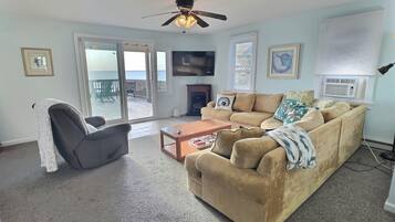 Ein Wohnzimmer mit einem beigen Sofa, einem Couchtisch, einem Sessel, einem Fernseher und einem Kamin. Es gibt einen Balkon mit Blick aufs Meer.