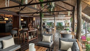 A spacious indoor area with wooden flooring, multiple seating arrangements including sofas and chairs, and a bar area with shelves of bottles. - La bastide de Caro (Siem Reap)