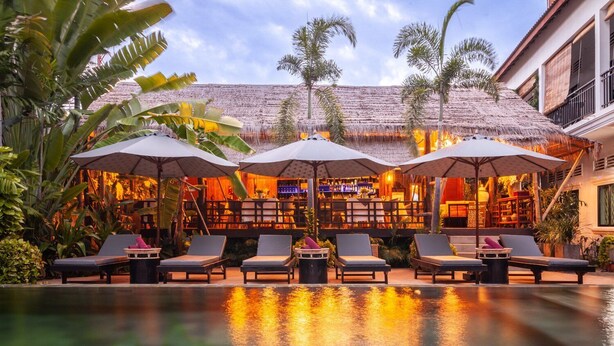 A poolside area with lounge chairs and umbrellas, a bar with a thatched roof, and tropical plants. - La bastide de Caro (Siem Reap)