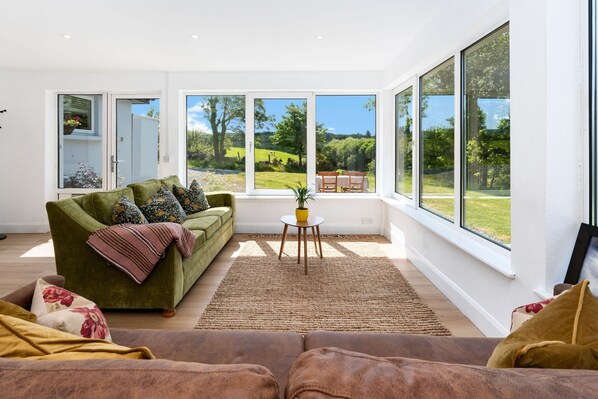 A modern living room with a green sofa, a brown sofa, and a small table with a plant. Large windows offer a view of a garden and a dining area.