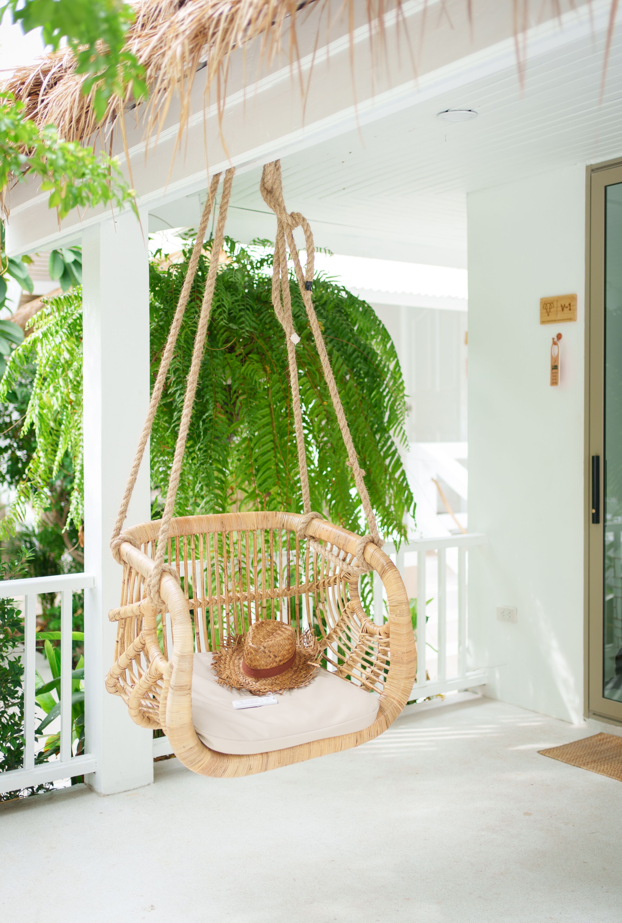 A wicker hanging chair with a cushion, a thatched roof, and lush greenery in the background.