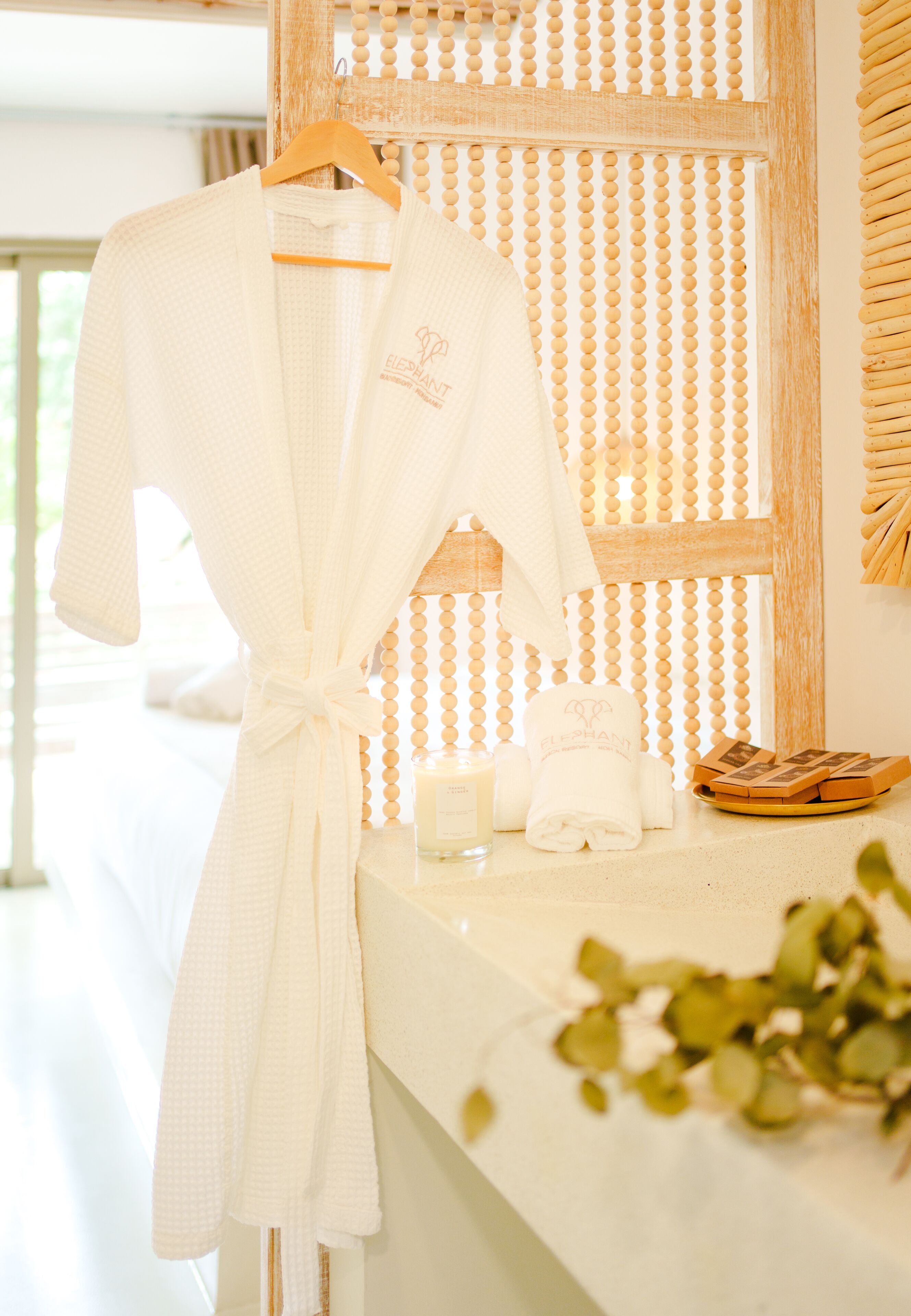 Une robe blanche pend à un cintre en bois, un ensemble de serviettes blanches est posé sur le comptoir de la salle de bain, ainsi qu’une bougie et un plateau contenant des produits de bain.