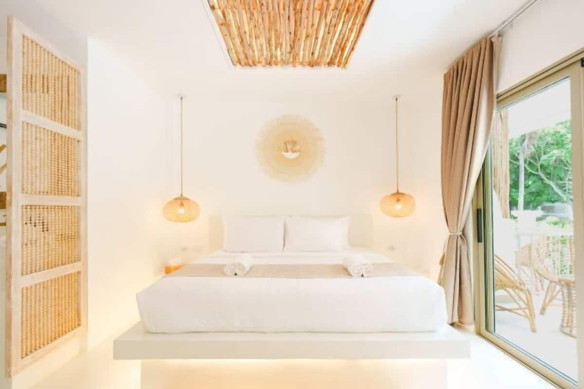 Un dormitorio moderno con una cama centrada sobre una plataforma blanca, dos lámparas colgantes, un ventilador de techo y vistas al exterior a través de un ventanal grande.