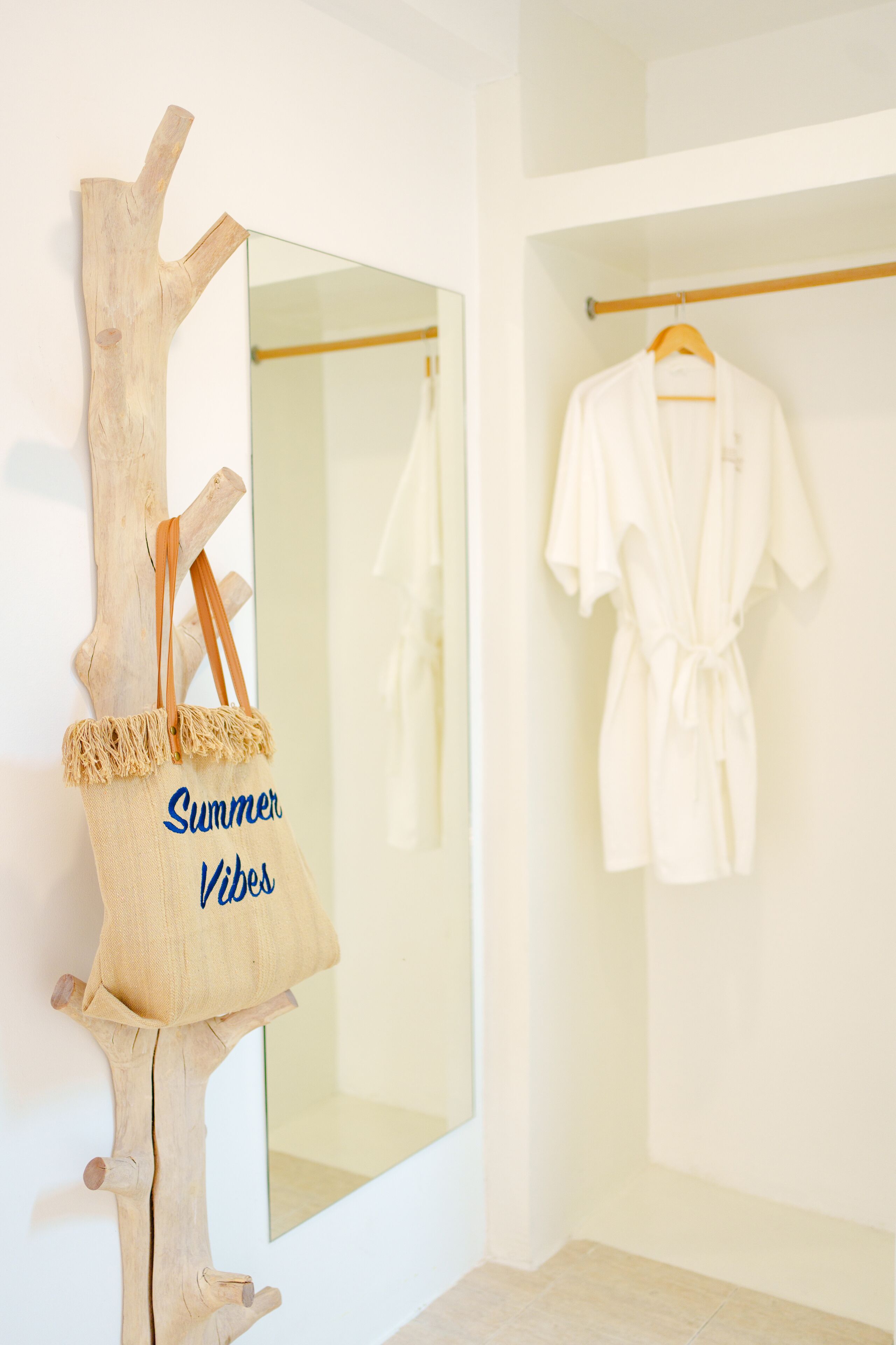 A wooden tree trunk with a canvas tote bag labeled "Summer Vibes" hanging from it, a white robe hanging on a rod, and a mirror reflecting the robe.