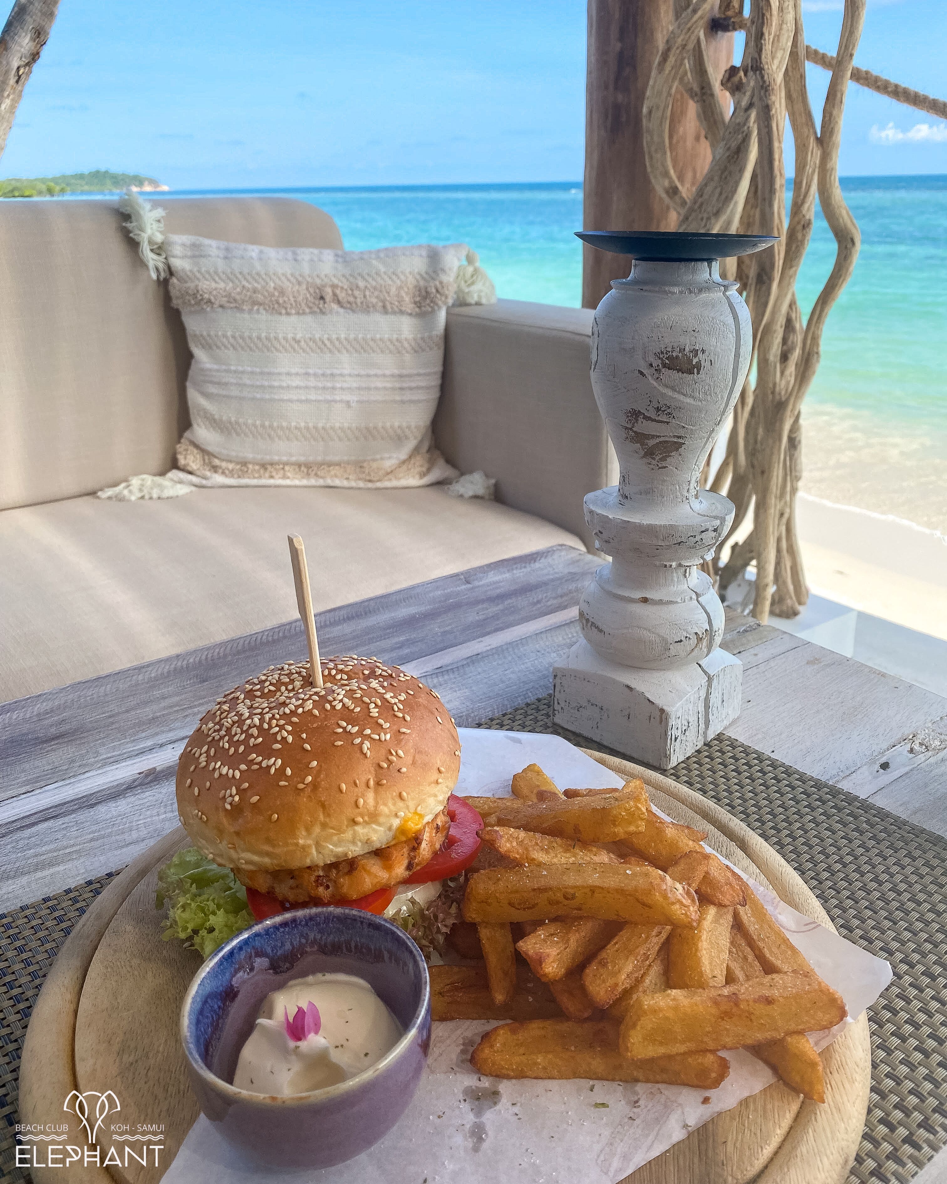 Une table dressée sur la plage, avec un hamburger, des frites et une sauce pour tremper, le tout présenté sur une planche en bois, avec vue sur l’océan.