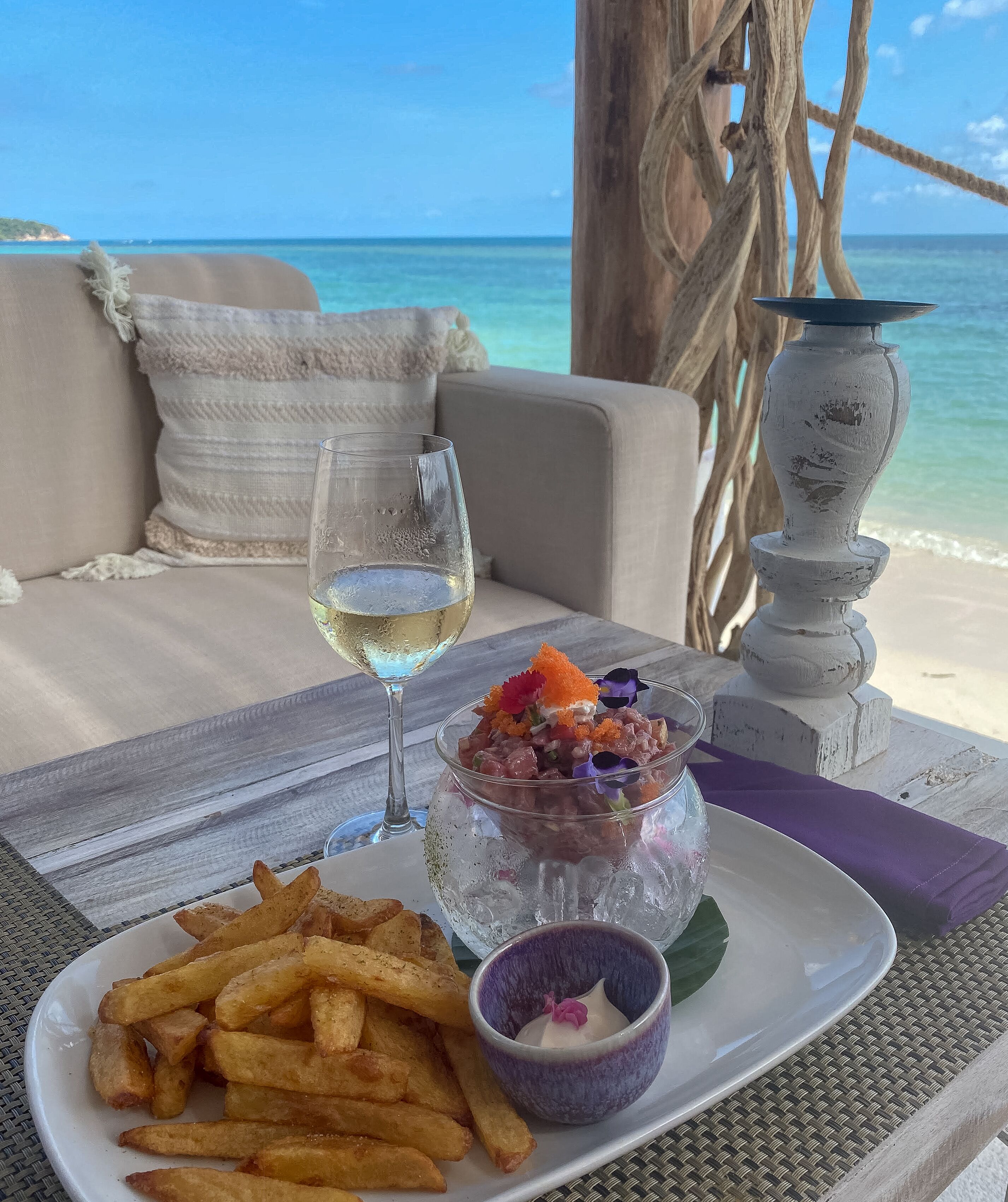 Ein Tisch mit einem Glas Weißwein, einer Schale Meeresfrüchtsalat, Pommes und einer kleinen Soße, auf einem Strand mit Blick aufs Meer.