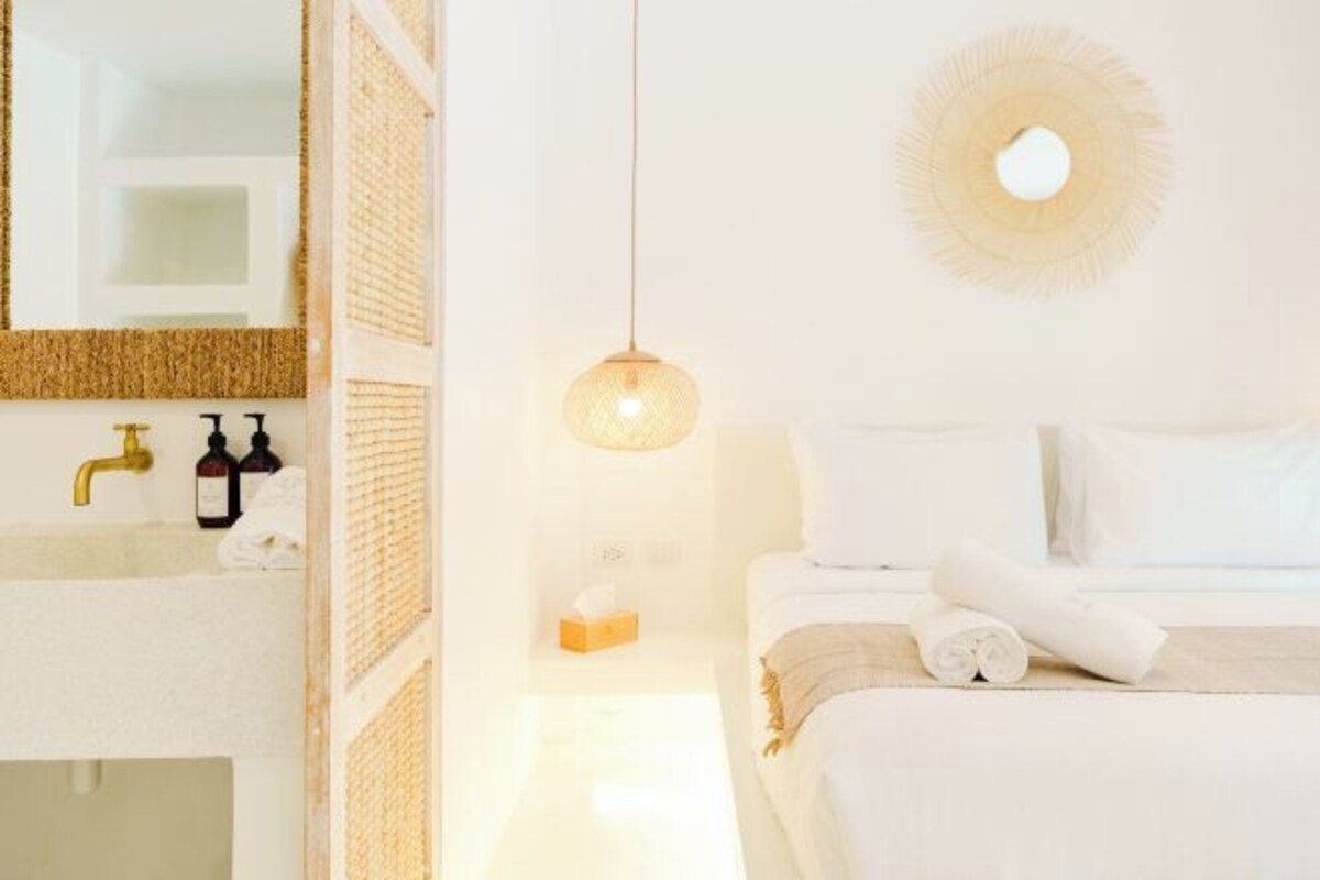 Habitación de hotel con cama blanca, espejo redondo en la pared, lavamanos blanco con grifos dorados y lámpara colgante.