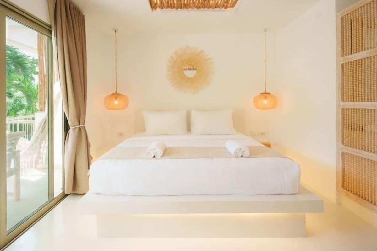 A bed with white linens, two bedside lamps, a sunburst wall decoration, and a view of greenery outside.