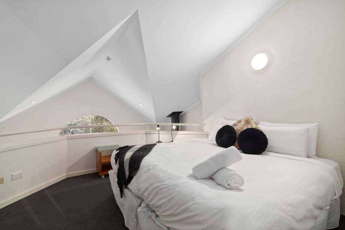 Een slaapkamer met een schuine kap, een bed met witte beddengoed, zwarte kussens en een nachtkastje met een lamp.