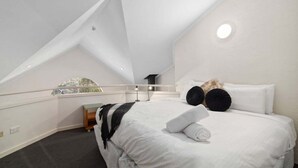 A bedroom with a sloped ceiling, a bed with white bedding, black pillows, and a bedside table with a lamp. - Inala 2 is an exceptional top floor, one bedroom and loft apartment. (Thredbo)