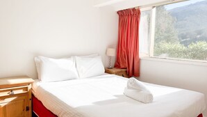 A neatly made bed with white linens, a wooden nightstand with a lamp, and a window with red curtains.