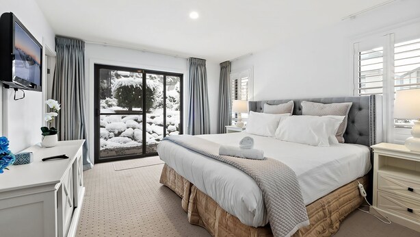Een moderne slaapkamer met een groot bed, een aan de muur bevestigde televisie, een schuifdeur naar een besneeuwd terras, en een nachtkastje met een lamp.