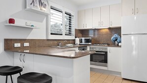 Een moderne keuken met witte kasten, een granieten aanrecht, een roestvrijstalen fornuis en een koelkast. Er staan twee zwarte barkrukken bij en er is een raam met zonwering.