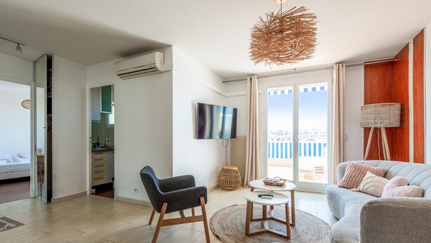 A modern living room with a sofa, armchair, and ottoman, a television, and a view of the outdoors. - La Terrasse Zéphyr - Welkeys (Antibes)