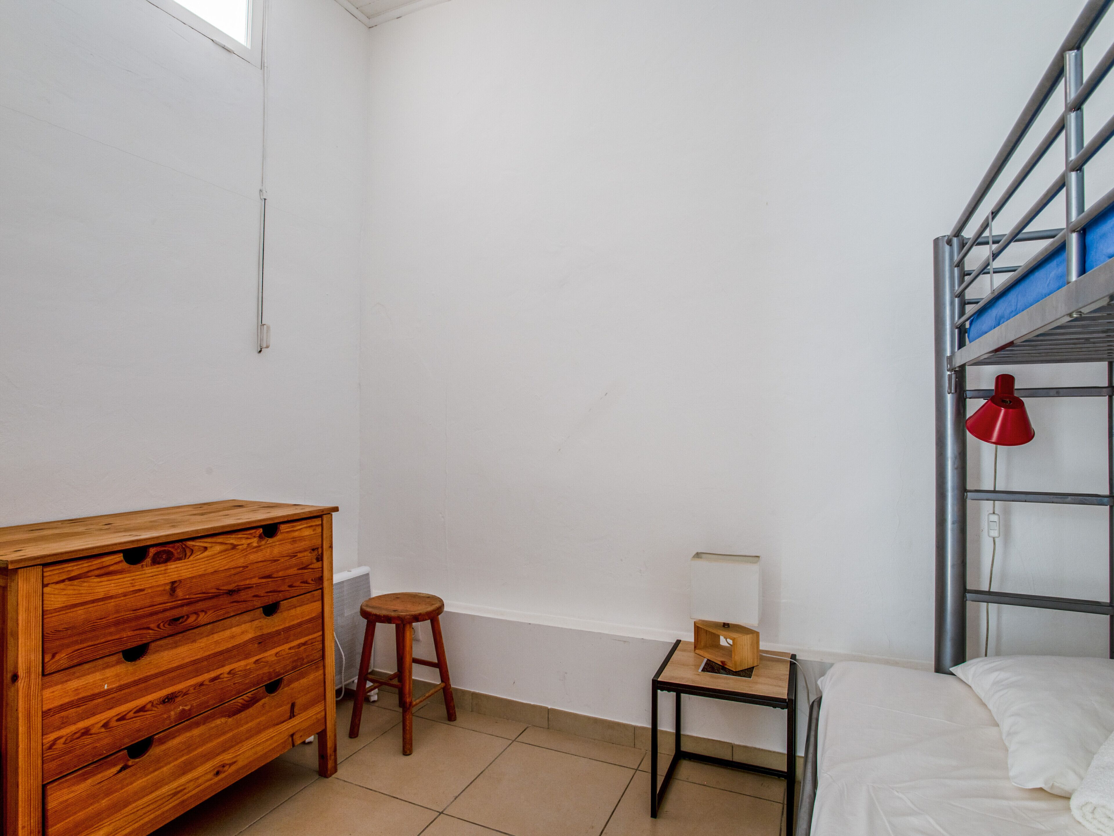 A room with a wooden dresser, a stool, a small table with a lamp, a bunk bed with a red lamp, and a radiator.