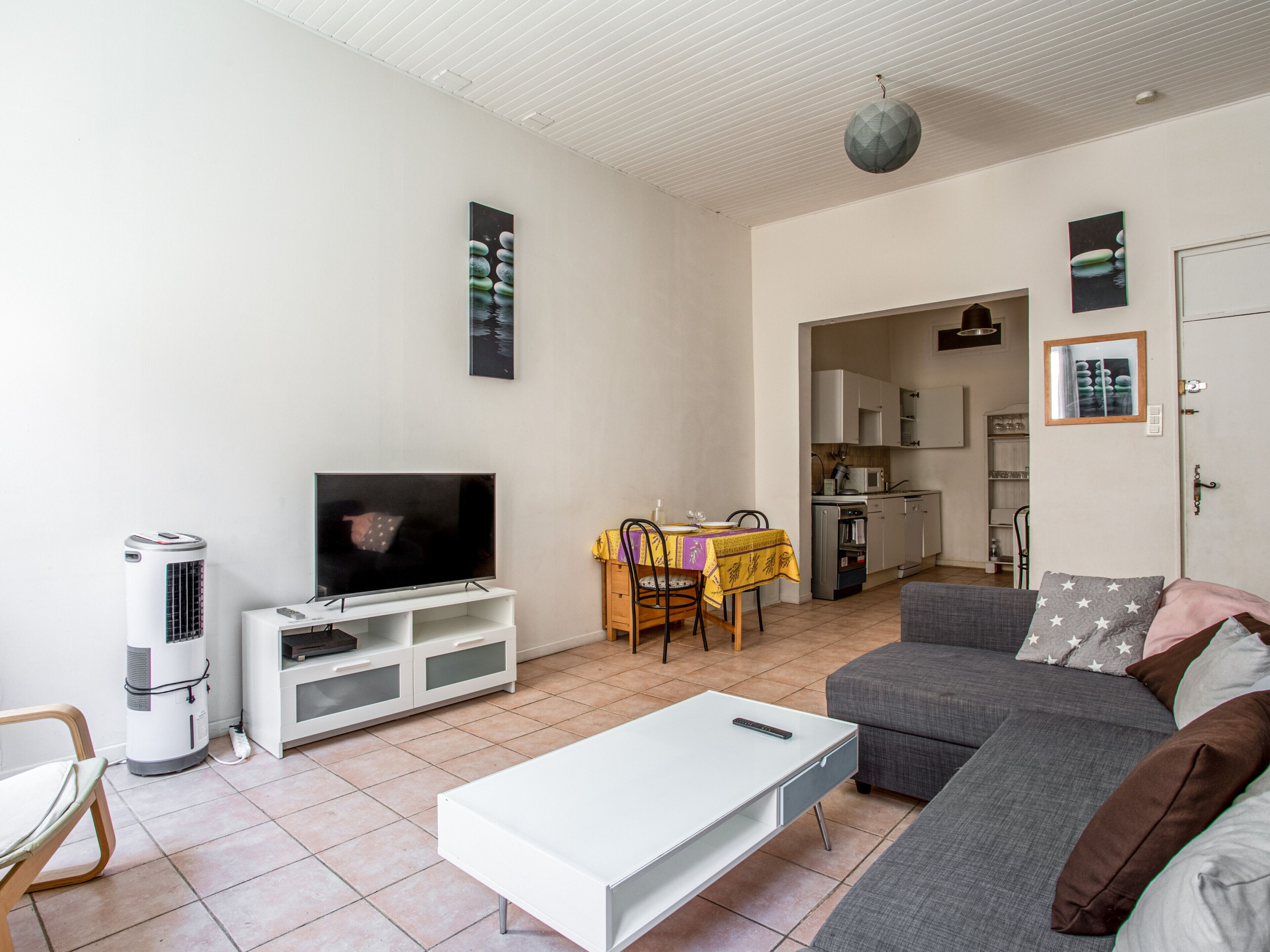 A living room with a flat-screen TV, a white coffee table, a gray sofa, and a dining area with a yellow tablecloth.