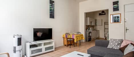 A living room with a flat-screen TV, a white coffee table, a gray sofa, and a dining area with a yellow tablecloth.