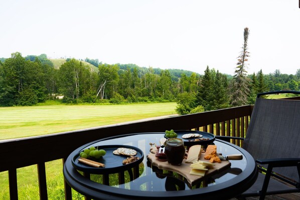 A table with cheese, crackers, and grapes on a balcony overlooking a grassy field and forested hills.