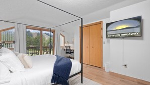A bedroom with a large bed, a TV displaying 'Skyview Hideaway', a wooden door, and a balcony with a view. - Summer Adventure Retreat - Golf Views, King Beds, Fully Loaded Kitchen! (Mancelona)