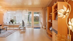Ein Zimmer mit Holzbalkendecke, einem Schreibtisch mit Computer, einem Stuhl, einem Bücherregal und einem großen Fenster mit Vorhängen.