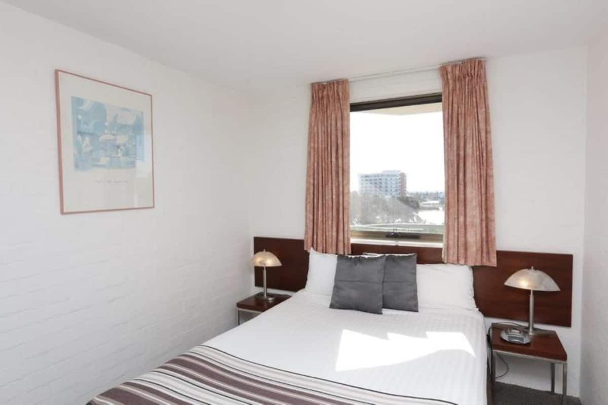 A hotel room with a large bed, two bedside tables with lamps, a window with curtains, and a framed picture on the wall.