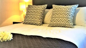 Un lit bien fait, agrémenté d’oreillers à motifs et d’une couverture sombre, une lampe de chevet et une fleur posée sur le lit.