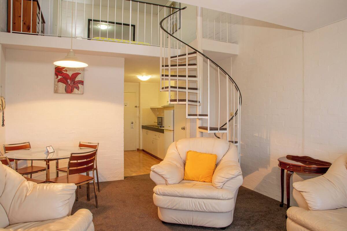 A living room with a white leather sofa, a dining table with chairs, a white armchair with a yellow pillow, and a spiral staircase leading to an upper floor.