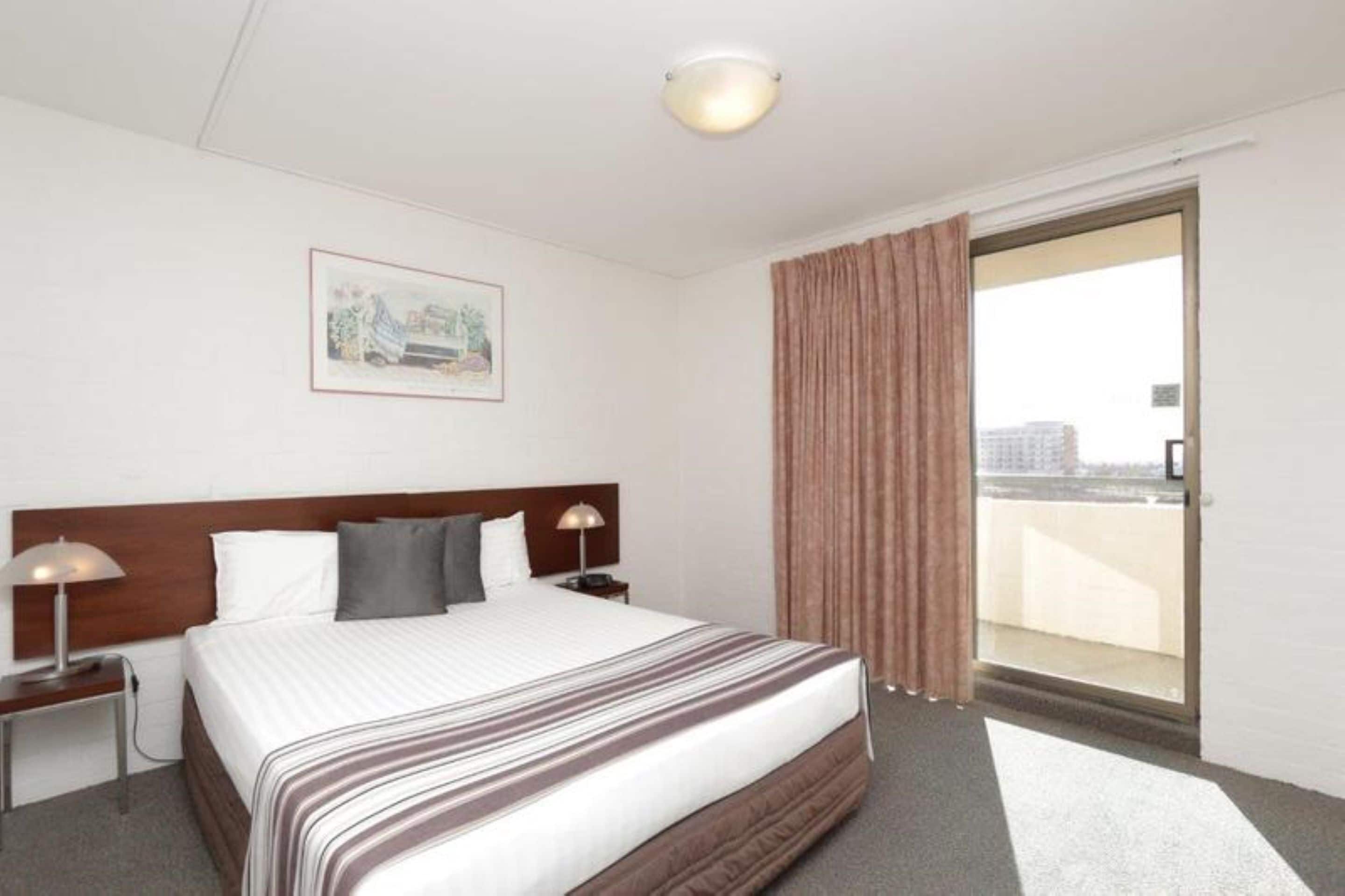 A hotel room with a large bed, a headboard, two bedside lamps, a nightstand, a framed picture on the wall, and a sliding glass door leading to a balcony.