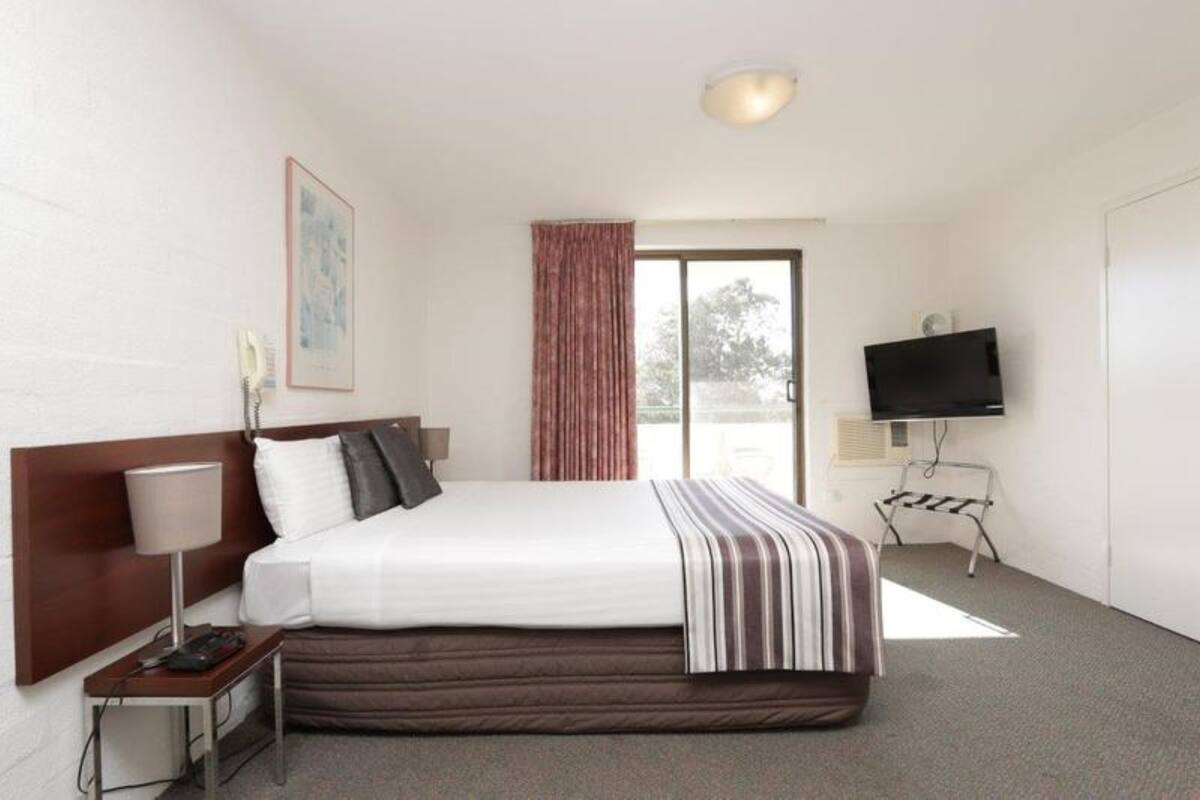 A hotel room with a bed, a TV, a lamp, a painting, and a window with curtains.