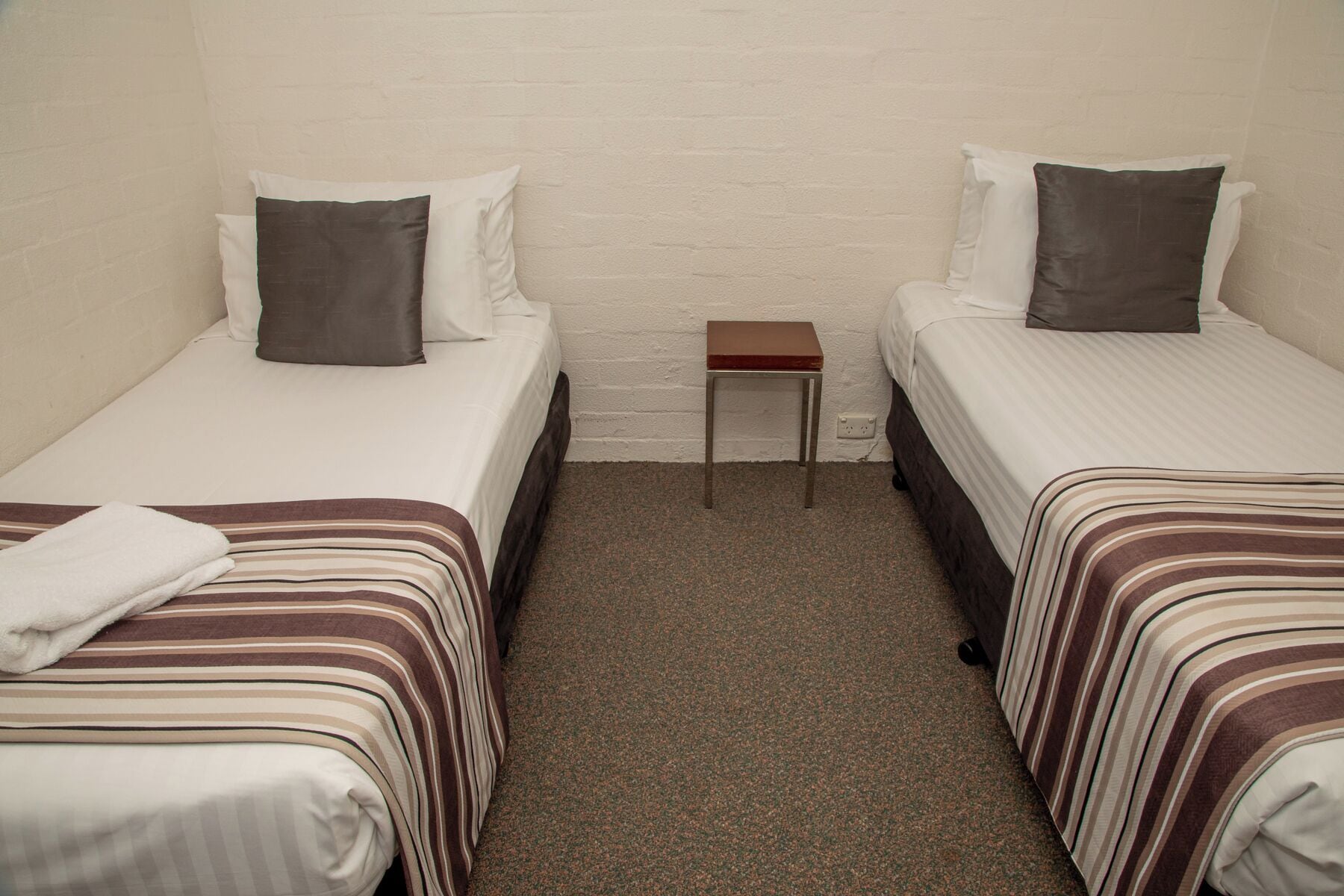 A small, clean room with two single beds, a small table, and a white brick wall.