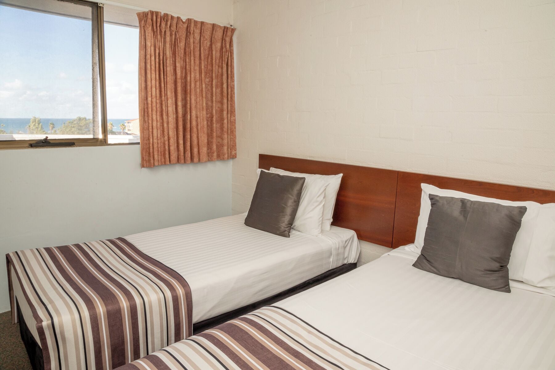 A hotel room with two single beds, a wooden headboard, a window with curtains, and a view of the sea.