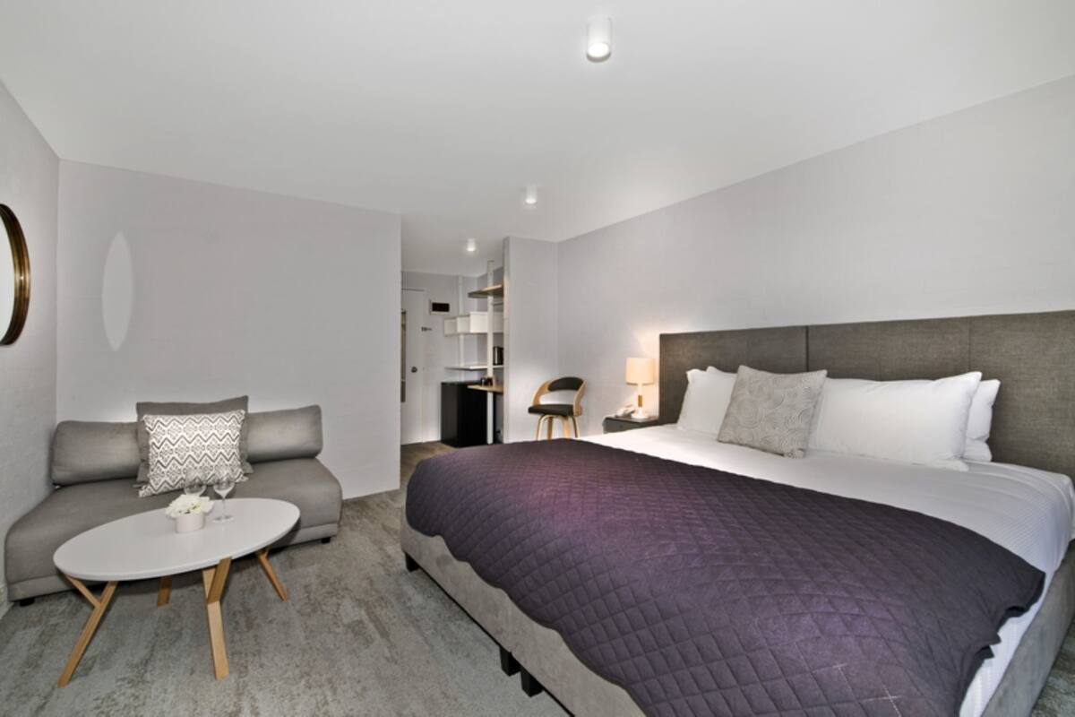 A modern hotel room with a large bed, a sofa, a small round table, and a desk with a lamp.