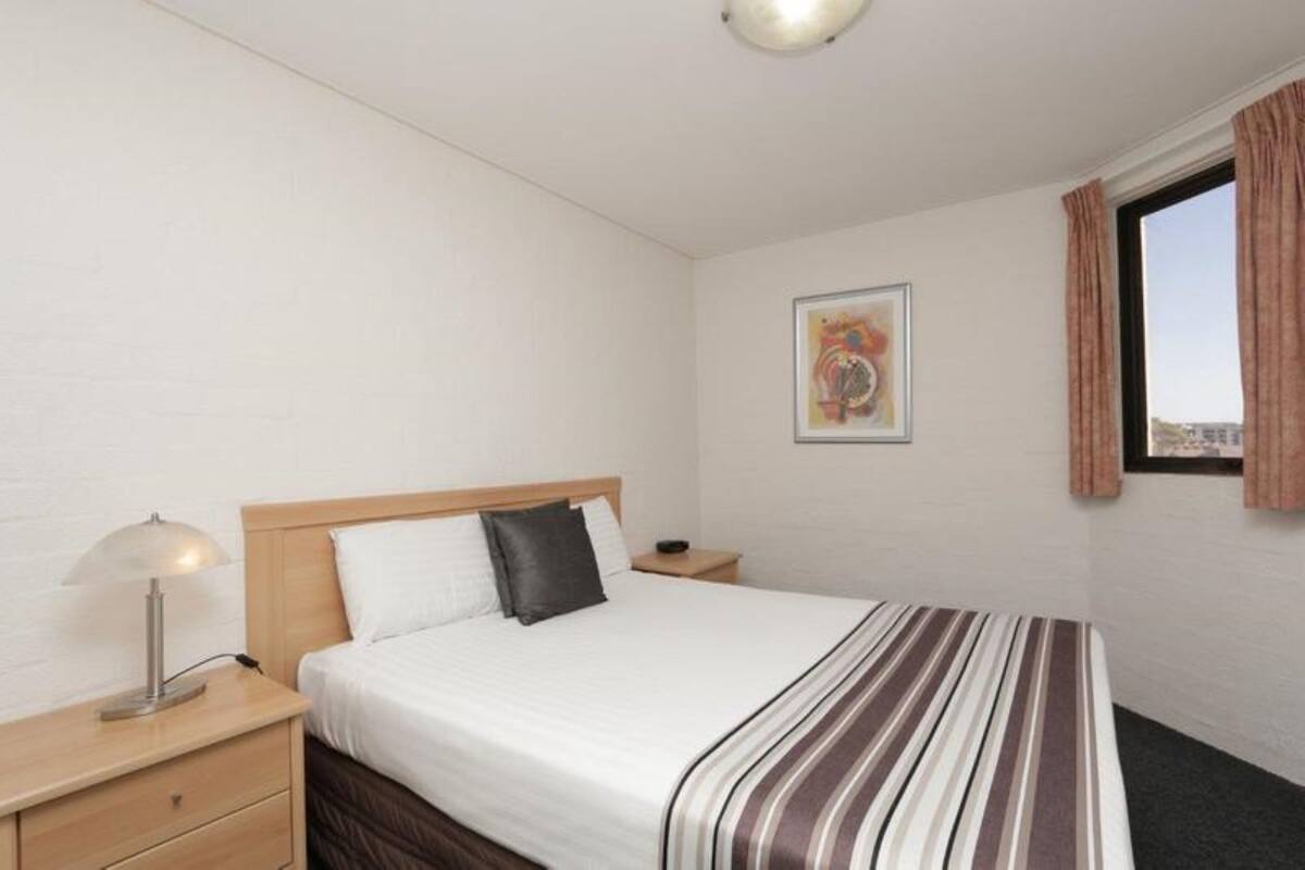 A hotel room with a bed, a nightstand, a lamp, a painting on the wall, and a window with curtains.
