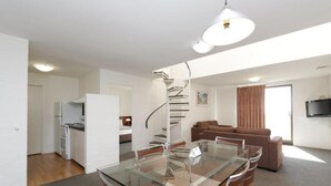 Un appartement moderne doté d’une table à manger en verre, d’une cuisine avec des meubles blancs, d’un escalier en colimaçon, d’un salon avec un canapé marron et d’un téléviseur à écran plat fixé au mur.
