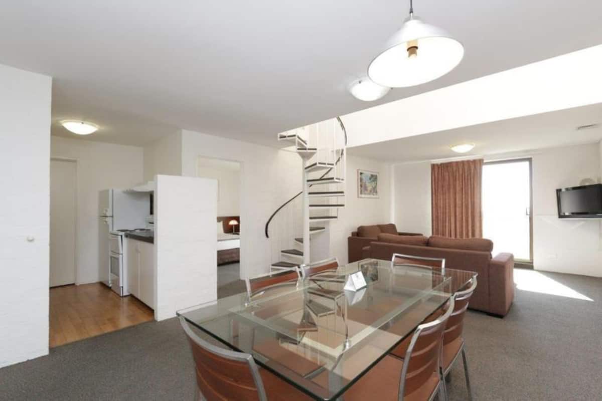 A modern apartment with a glass dining table, a kitchen with white cabinets, a spiral staircase, a living area with a brown sofa, and a flat-screen TV mounted on the wall.