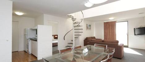 A modern apartment with a glass dining table, a kitchen with white cabinets, a spiral staircase, a living area with a brown sofa, and a flat-screen TV mounted on the wall.